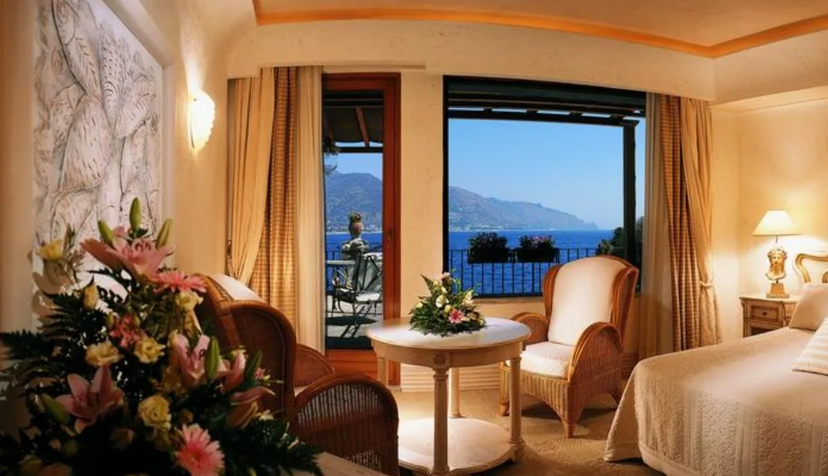 Camera elegante con letto, poltrone in vimini e balcone con vista mare